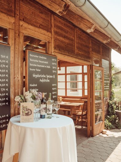 A cozy wooden cottage with a table and a flower vase decoration. Boards with menu offers are hanging on the wall. | © Weingut Kleinschuster