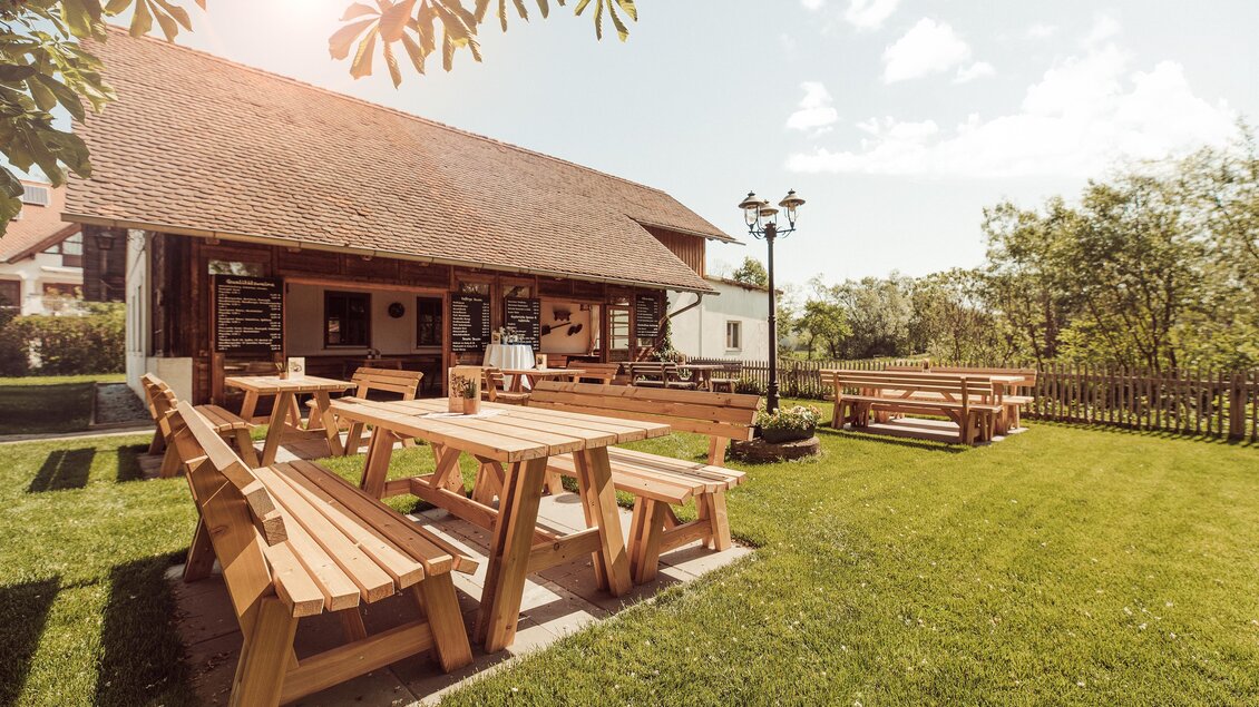 Ein einladender Garten mit Holzmöbeln und einem rustikalen Gebäude im Hintergrund. Die Atmosphäre ist freundlich und sonnig. | © Weingut Kleinschuster