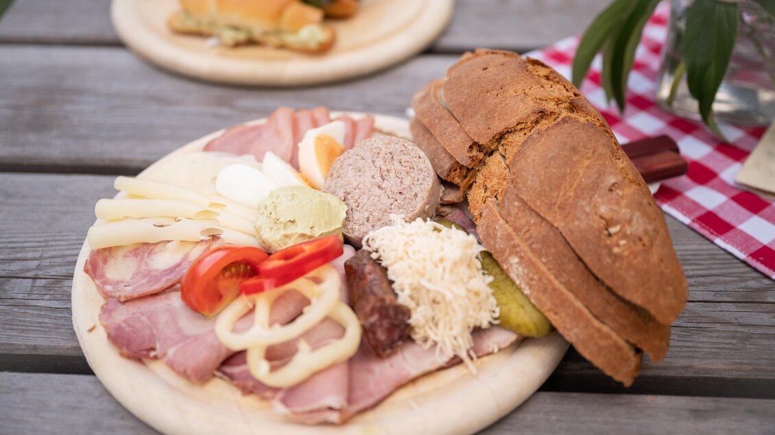 Ein Teller mit verschiedenen Aufschnittsorten, Käse und Gemüse, dazu ein Stück Roggenbrot. Der Hintergrund zeigt eine rustikale Holztischoberfläche. | © Weingut Kleinschuster
