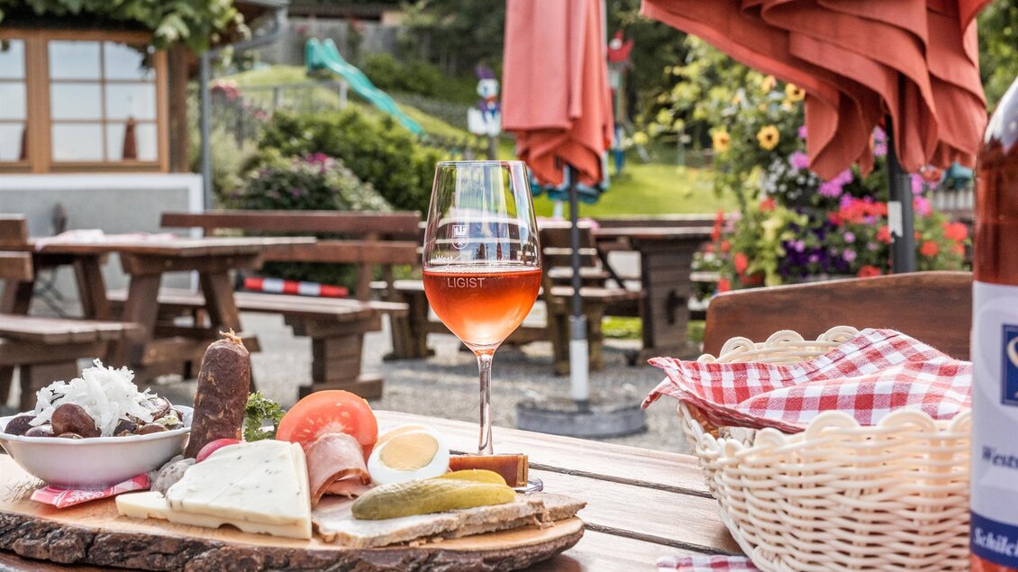 Ein gedeckter Tisch im Freien mit einer Vielzahl von regionalen Köstlichkeiten auf einem Holzbrett. Dazu ein Glas Roséwein und eine schöne Gartenlandschaft im Hintergrund. | © Die Abbilderei
