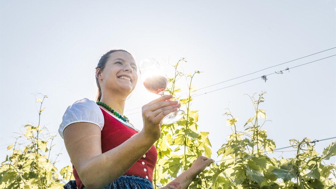 Eine Frau in traditioneller Kleidung steht in einem Weinberg und hält ein Glas Wein in der Hand. Die Sonne strahlt im Hintergrund und sorgt für eine fröhliche Atmosphäre. | © Die Abbilderei