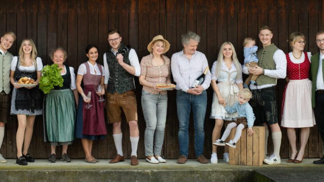 Eine Gruppe von Menschen in traditioneller Kleidung steht vor einer Holzverkleidung. Sie halten Gegenstände wie Essen und Getränke und scheinen fröhlich zu sein. | © HP Dorner