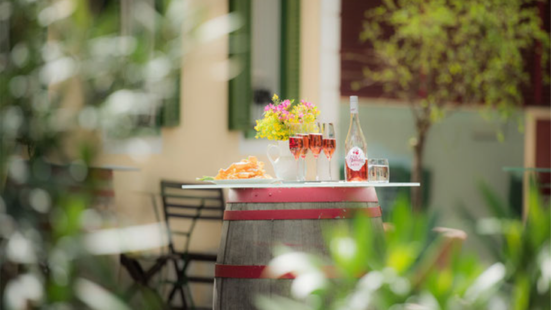 Ein Tisch aus einem Weinfass mit Cocktails und Snacks steht in einem Garten. Im Hintergrund sind grüne Pflanzen und Fenster zu sehen. | © HP Dorner