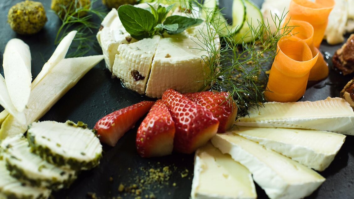 Eine kunstvoll arrangierte Käseplatte aus dem Buschenschank Dorner, mit verschiedenen Käsesorten, frischen Erdbeeren, Karottenstreifen und Zucchini. Dazu sind Kräuter und Pistazienbällchen dekorativ angerichtet. | © Strobl Christa