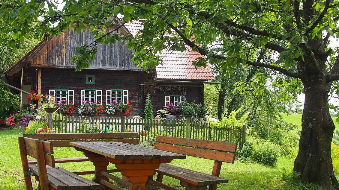 Ein traditionelles Holzhaus mit bunten Blumenfenstern. Vor dem Haus steht eine Holzbank unter einem Baum. | © Buschenschank Eckfastl