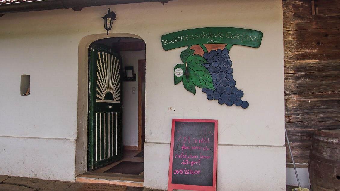 Ein gemütlicher Eingang zu einem Weingut mit einer bunten Traubenmalerei. Vor der Tür steht ein schwarzes Schild mit Informationen. | © Buschenschank Eckfastl