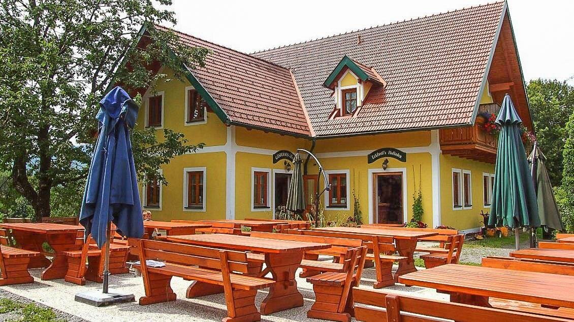 Ein charmantes gelbes Gebäude mit einem roten Dach und Holz-Terrassenmöbeln. Der Außenbereich ist mit sonnenschützenden Schirmen ausgestattet und von Bäumen umgeben. | © Buschenschank Eckfastl