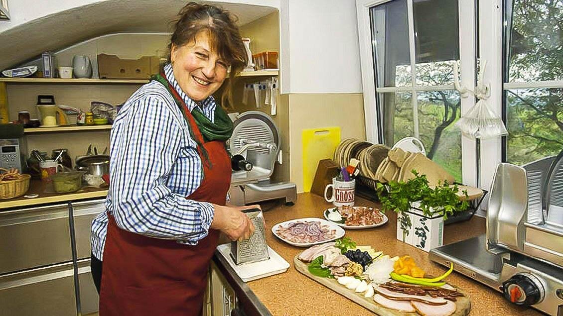 Eine Frau in einer Küche lächelt, während sie Fleisch und Gemüse für eine Mahlzeit vorbereitet. Auf der Arbeitsfläche liegt eine Auswahl an frischen Zutaten. | © Buschenschank Eckfastl