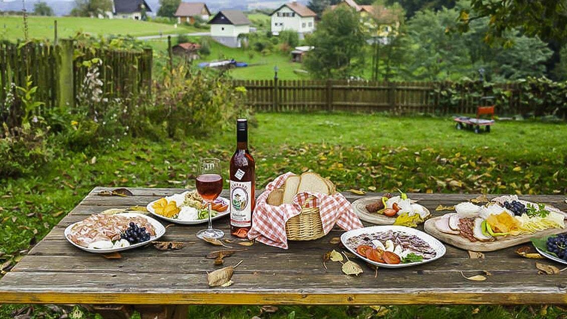 Ein schöner Tisch im Freien, gedeckt mit verschiedenen Speisen und Getränken. Im Hintergrund sind grüne Wiesen und gemütliche Häuser zu sehen. | © Buschenschank Eckfastl