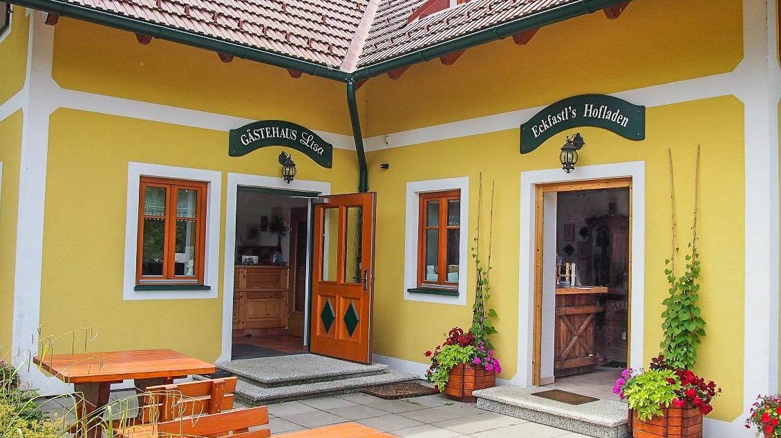 Ein charmantes Gästehaus mit gelber Fassade und zwei Eingängen. Vor dem Gebäude stehen Tische und Stühle, umgeben von bunten Pflanzen. | © Buschenschank Eckfastl