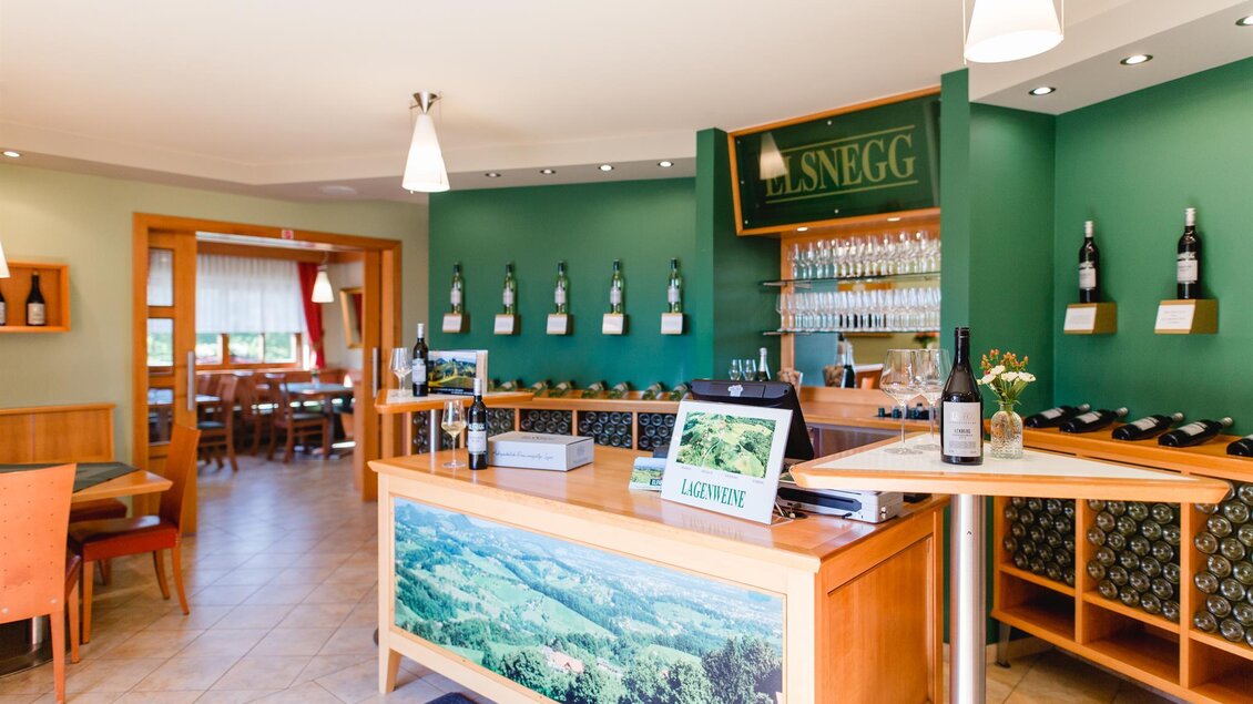 Ein einladendes Restaurant mit warmen Farben und einer Bar, die mit Weinen dekoriert ist. Der Raum ist hell und freundlich mit komfortablen Sitzgelegenheiten. | © Weingut Elsnegg