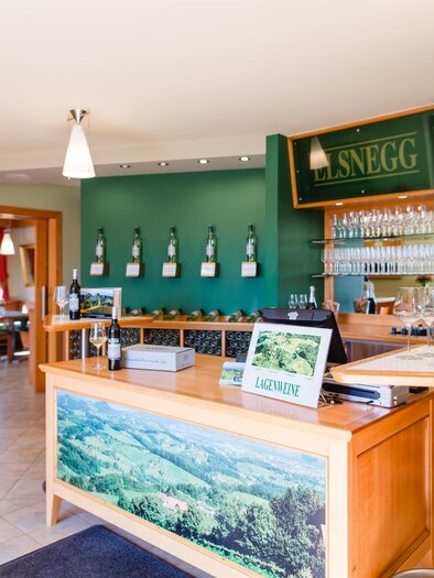 Ein einladendes Restaurant mit warmen Farben und einer Bar, die mit Weinen dekoriert ist. Der Raum ist hell und freundlich mit komfortablen Sitzgelegenheiten. | © Weingut Elsnegg