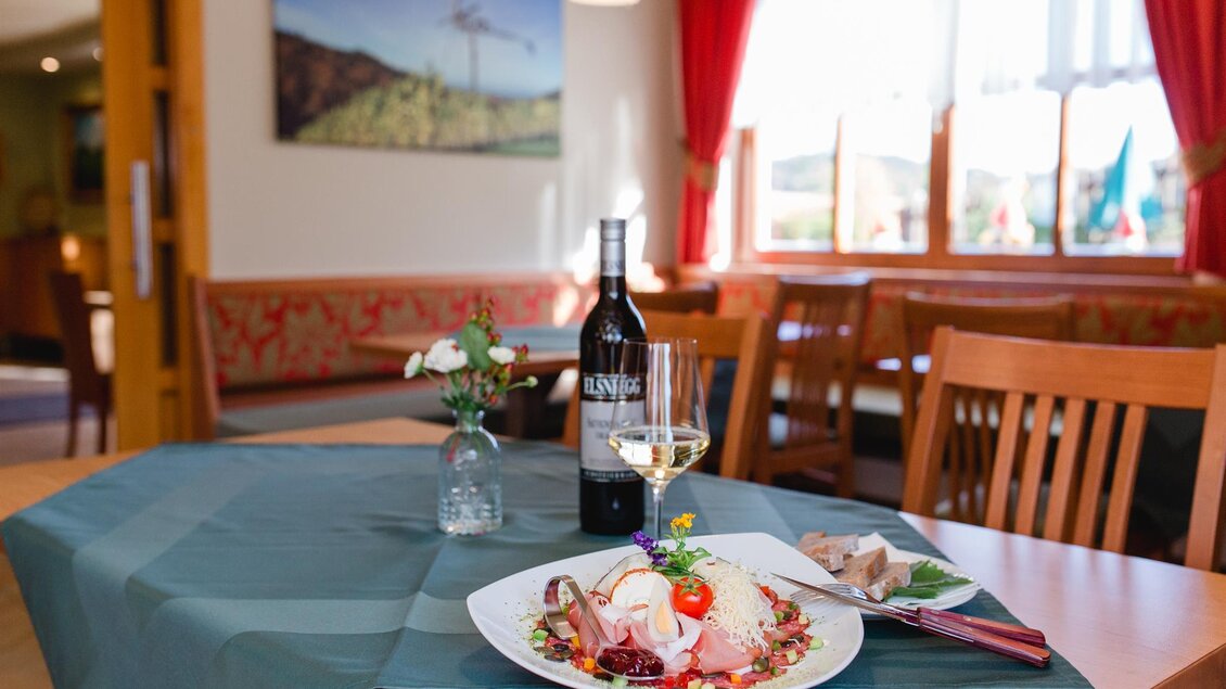 Ein schön gedeckter Tisch in einem Restaurant mit einem Gericht und einem Glas Weißwein. Im Hintergrund sind gemütliche Sitzgelegenheiten und große Fenster zu sehen. | © Weingut Elsnegg