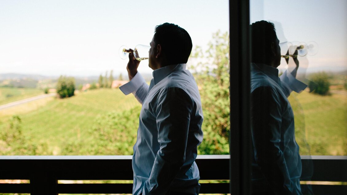 Ein Mann in einem weißen Hemd steht auf einem Balkon und schaut nach draußen. Er hält ein Glas in der Hand und genießt die Aussicht auf die grüne Landschaft. | © Weingut Elsnegg