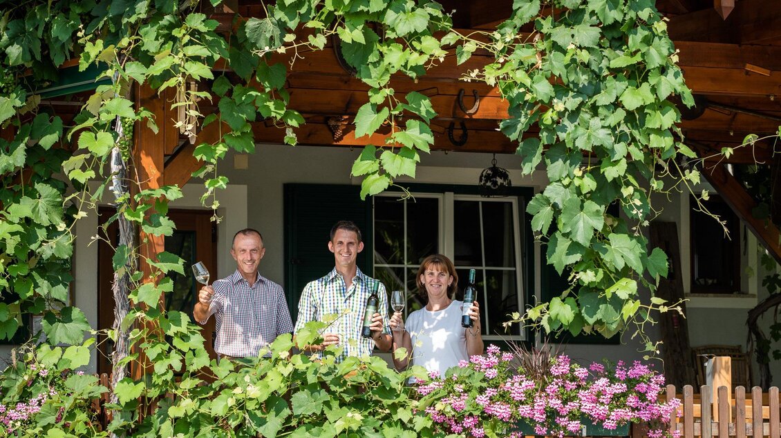 Ein herzliches Trio steht unter einem mit grünen Blättern bewachsenen Vordach. Sie halten Gläser in der Hand und lächeln freundlich. | © TVB Bad Waltersdorf