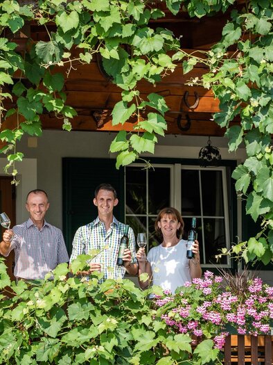 A warm trio stands under a canopy covered with green leaves. They are holding glasses in their hands and smiling friendly. | © TVB Bad Waltersdorf
