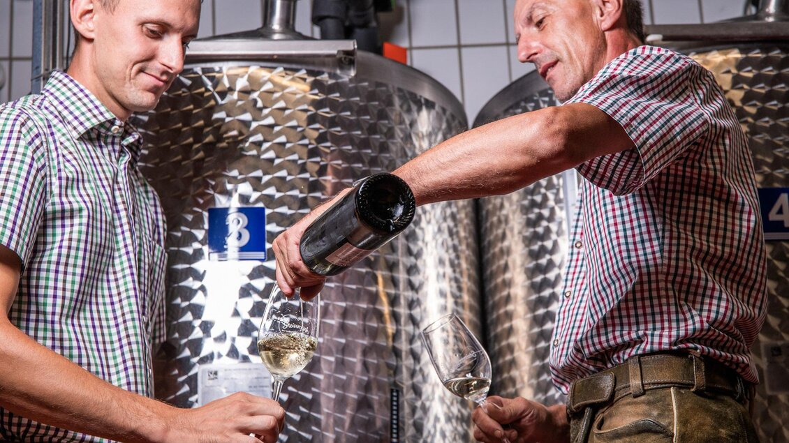 Zwei Männer stehen in einer Weinlagerung und gießen Wein in Gläser. Im Hintergrund sind große metalne Fässer zu sehen. | © TVB Bad Waltersdorf