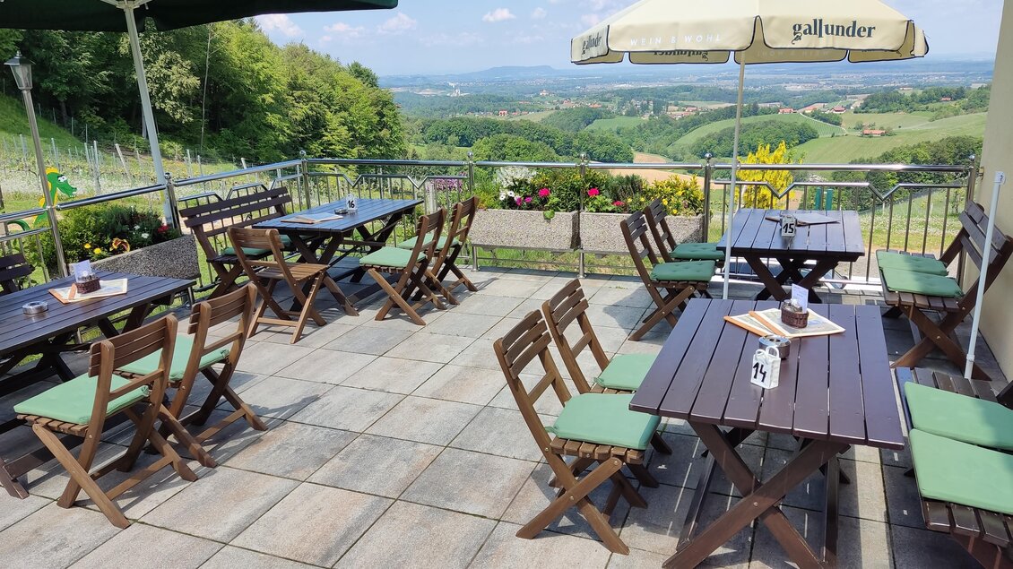 Eine gemütliche Terrasse mit Holztischen und Stühlen, umgeben von grünen Hügeln. Zwei Sonnenschirme spenden Schatten an einem sonnigen Tag. | © Sabine Gallunder