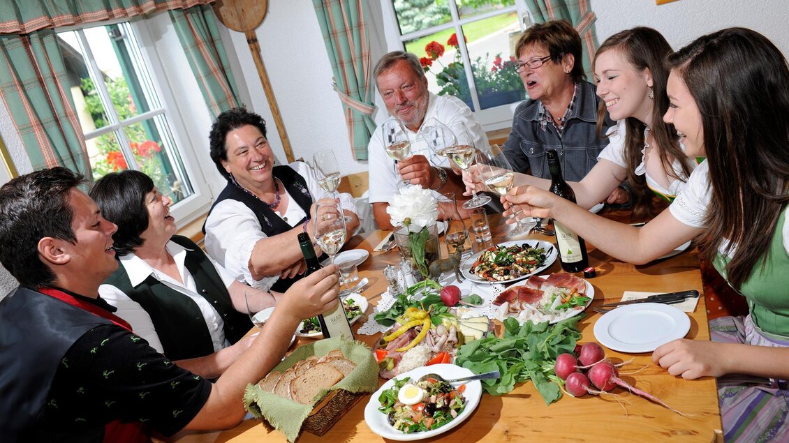 Eine gesellige Gruppe von Menschen sitzt an einem Tisch und hebt Gläser. Der Tisch ist reich gedeckt mit verschiedenen Speisen und frischen Zutaten. | © Weingut Gerngross