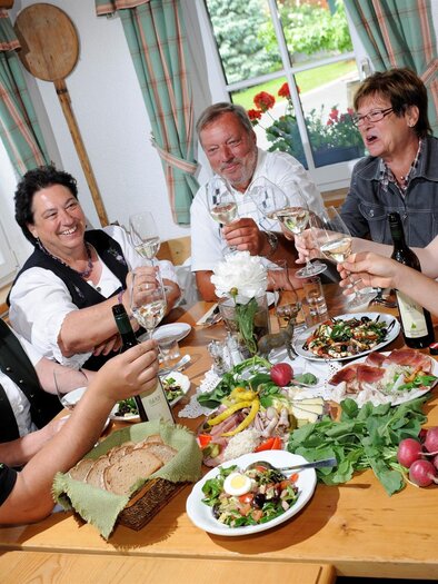 A sociable group of people is sitting at a table and raising their glasses. The table is richly laid with various dishes and fresh ingredients. | © Weingut Gerngross
