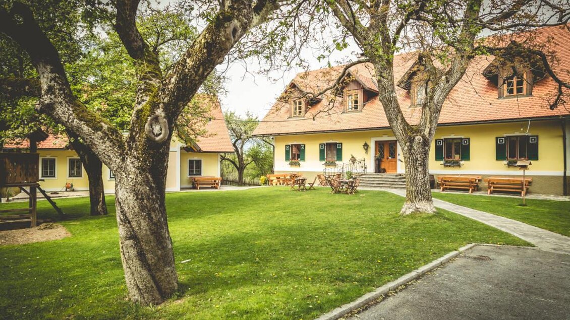 Ein gemütliches Bauernhaus mit rotem Dach in einem grünen Garten. Alte Bäume und Bänke laden zum Entspannen ein. | © Buschenschank Glirsch