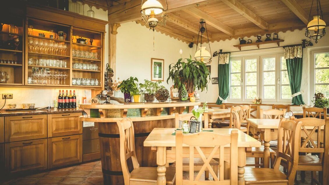 Ein gemütliches Restaurant mit Holzmöbeln und einer einladenden Bar. Der Raum ist hell erleuchtet und hat Fenster mit grünen Vorhängen. | © Buschenschank Glirsch
