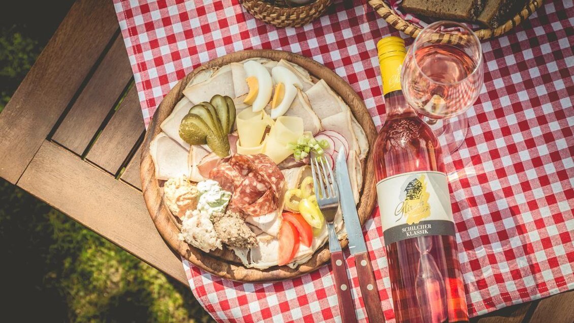 Ein köstlicher Teller mit Aufschnitt, Käse und Gemüse auf einem rot-weiß karierten Tischdecke. Daneben steht eine Flasche Roséwein und ein Glas. | © Buschenschank Glirsch
