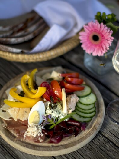 Ein schöner Teller mit verschiedenen Käsesorten, Wurst und frischem Gemüse. Daneben steht ein Glas Wein und eine Blume in einer Vase. | © Tom Lamm