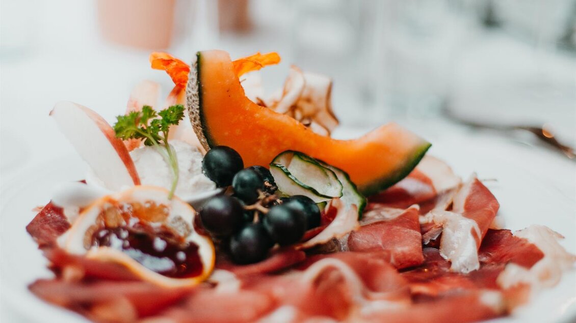 Ein appetitlicher Teller mit einer Auswahl an Schinken, frischen Früchten und einer dekorativen Garnitur. Der Hintergrund zeigt Gläser und einen festlichen Tisch. | © Weingut Gutjahr