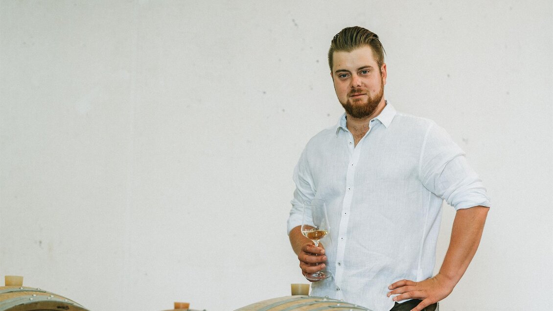 Ein Mann steht neben einem Weinfass und hält ein Glas in der Hand. Er trägt ein weißes Hemd und lächelt selbstbewusst. | © Weingut Gutjahr
