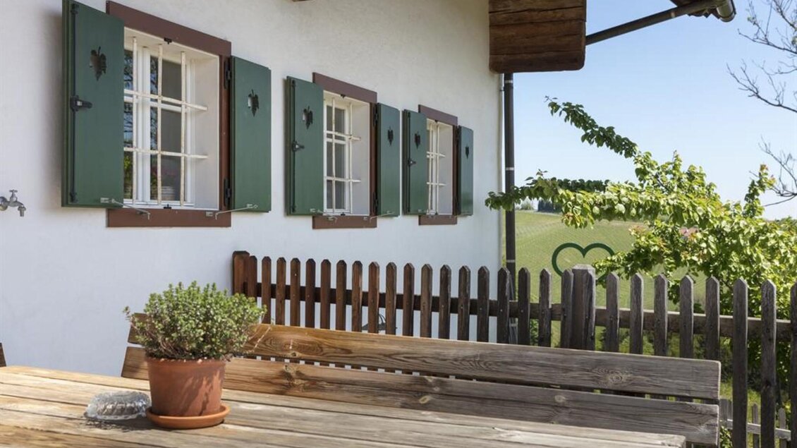 Ein gemütlicher Außenbereich mit einem Holztisch und einer Pflanze. Im Hintergrund sind Fenster mit grünen Fensterläden und eine schöne Landschaft zu sehen. | © Hack-Gebell