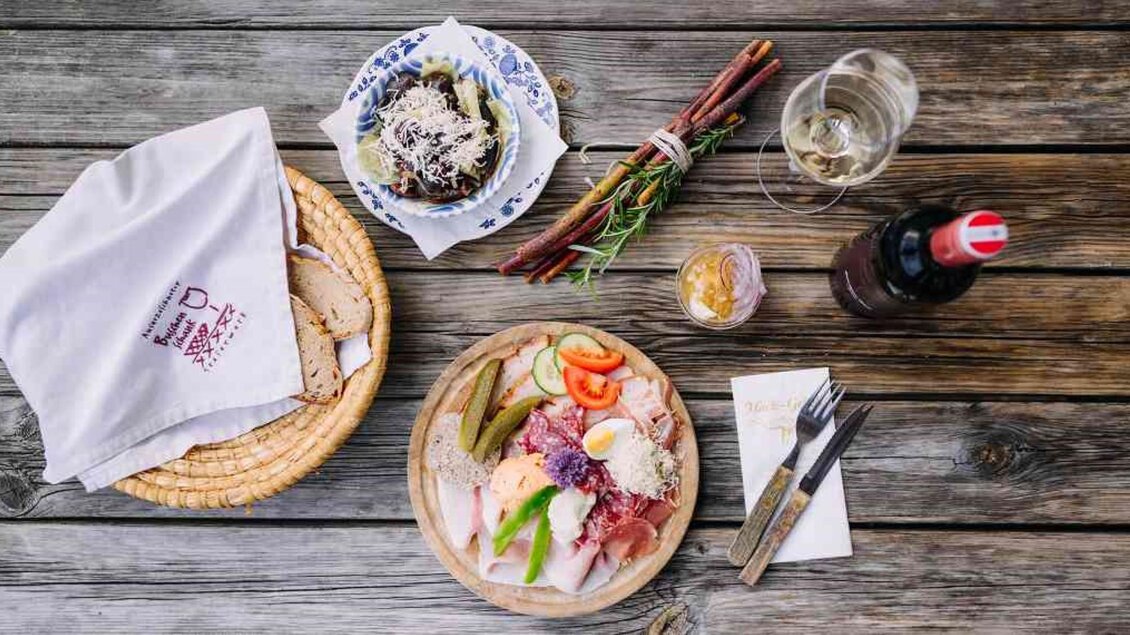 Ein Tisch mit mehreren Gerichten, darunter eine Platte mit Aufschnitt, frischem Gemüse und Brot. Neben einem Getränk sind auch einige Snacks und ein dekorierter Teller zu sehen. | © Hack-Gebell