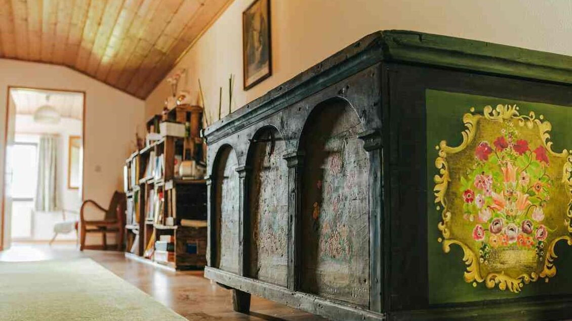 Ein traditioneller Schrank mit bunten Blumenmustern steht in einem stilvollen Raum. Im Hintergrund sieht man eine Bücherregal und einen gemütlichen Sessel. | © Hack-Gebell