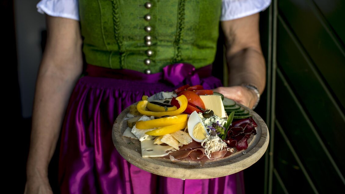 Eine Person trägt ein Holzbrett mit einer Auswahl an Wurst, Käse und Gemüse. Sie ist in einem traditionellen Outfit mit einer grünen Bluse und einem rosa Rock gekleidet. | © Hack-Gebell