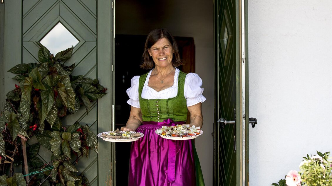Eine lächelnde Frau in traditioneller Tracht steht vor der Tür und hält zwei Tabletts mit Gebäck. Der Eingang ist von Pflanzen und Blumen umgeben. | © Hack-Gebell