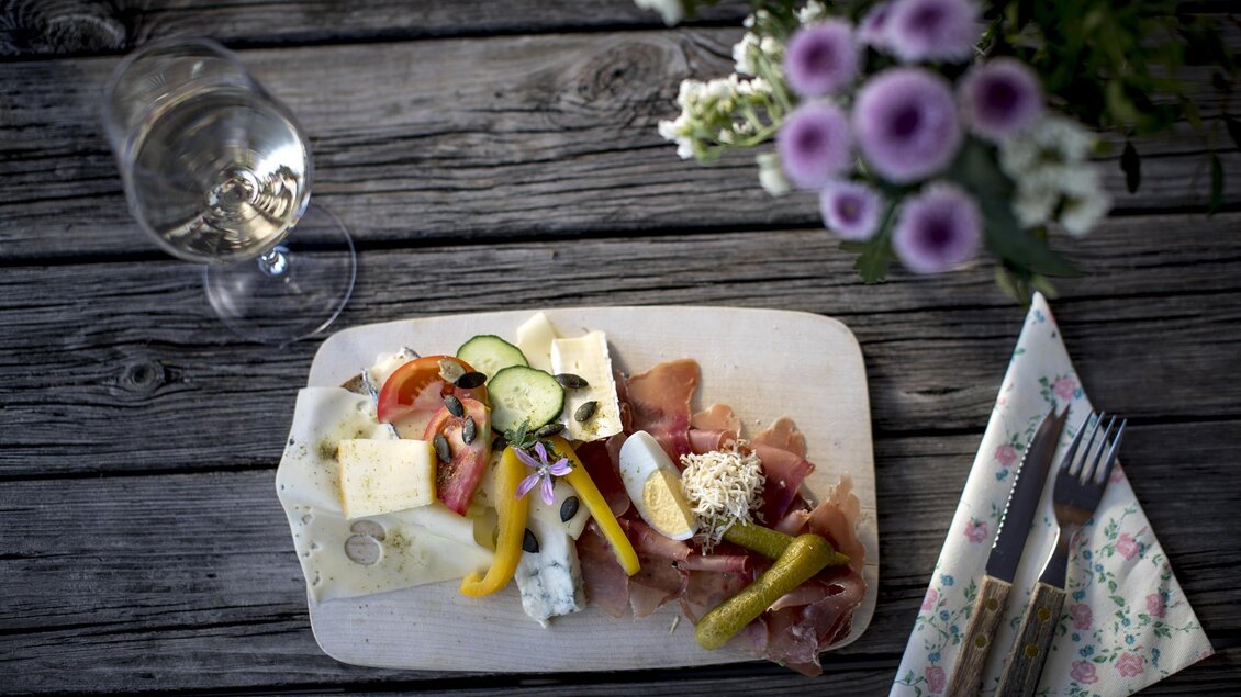 Ein rustikales Holzbrett mit einer Auswahl an Käse, Schinken und frischem Gemüse. Neben dem Brett steht ein Glas Wasser und ein Blumenstrauß. | © Tomm Lamm