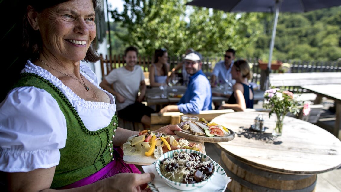 Eine freundliche Kellnerin trägt zwei Teller mit köstlichem Essen. Im Hintergrund sind Gäste an einem Tisch im Freien zu sehen. | © Tomm Lamm