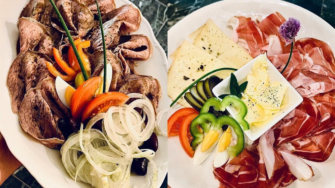 Ein Teller mit zartem Fleisch, frischen Zwiebeln und buntem Gemüse. Daneben ein weiterer Teller mit Käse und Aufschnitt, garniert mit Kräutern. | © Buschenschank Pichlippi