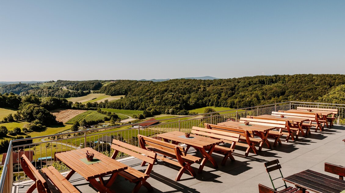 Eine sonnige Terrasse mit vielen Holztischen und Bänken. Im Hintergrund erstreckt sich eine grüne Landschaft unter einem klaren blauen Himmel. | © Familie Hartinger