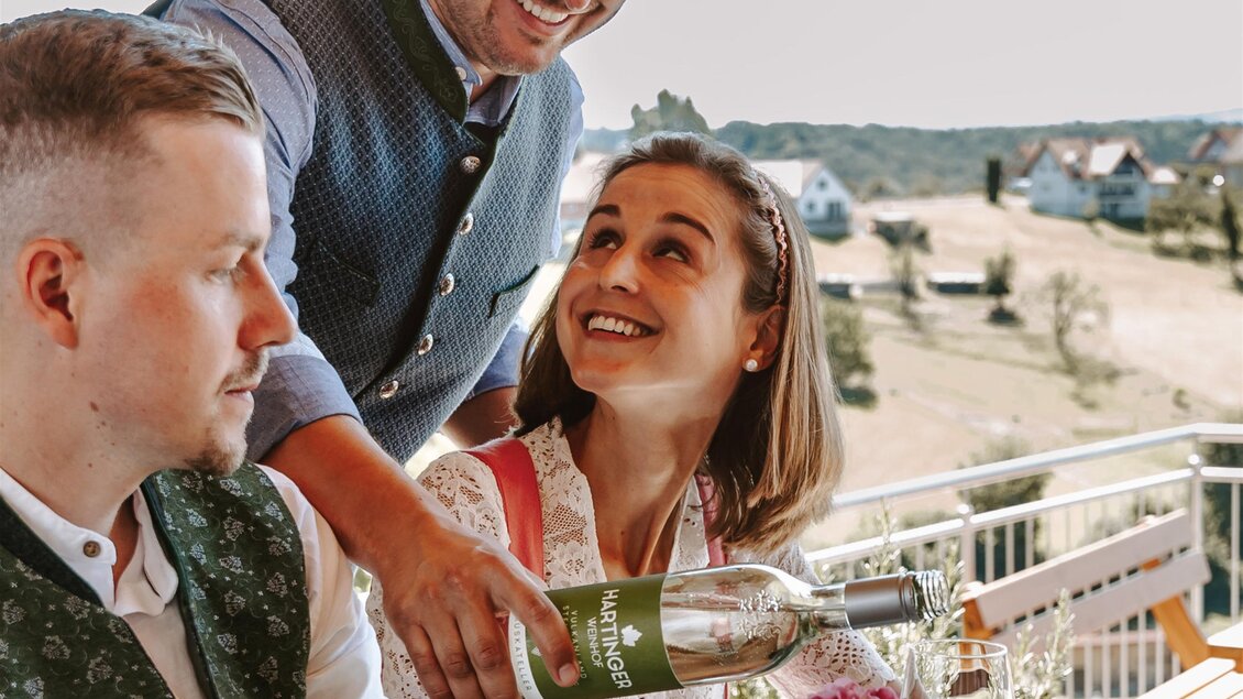 Ein freundlicher Kellner schenkt Wein in ein Glas für eine lächelnde Frau. Am Tisch sitzen zwei Personen in traditioneller Kleidung, umgeben von einer schönen Aussicht. | © Familie Hartinger