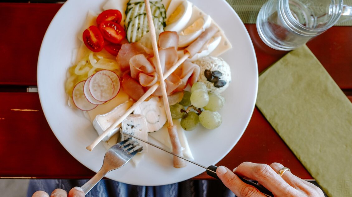 Ein Teller mit verschiedenen Käsesorten, Aufschnitt und frischem Gemüse. Dazu ein Glas Weißwein auf dem Tisch. | © Familie Hartinger