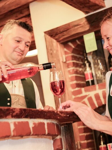 Ein Mann schenkt einer Frau ein Glas Roséwein hinter einer Bar ein. Beide tragen traditionelle Trachten und lächeln freundlich. | © Buschenschank Haubensima