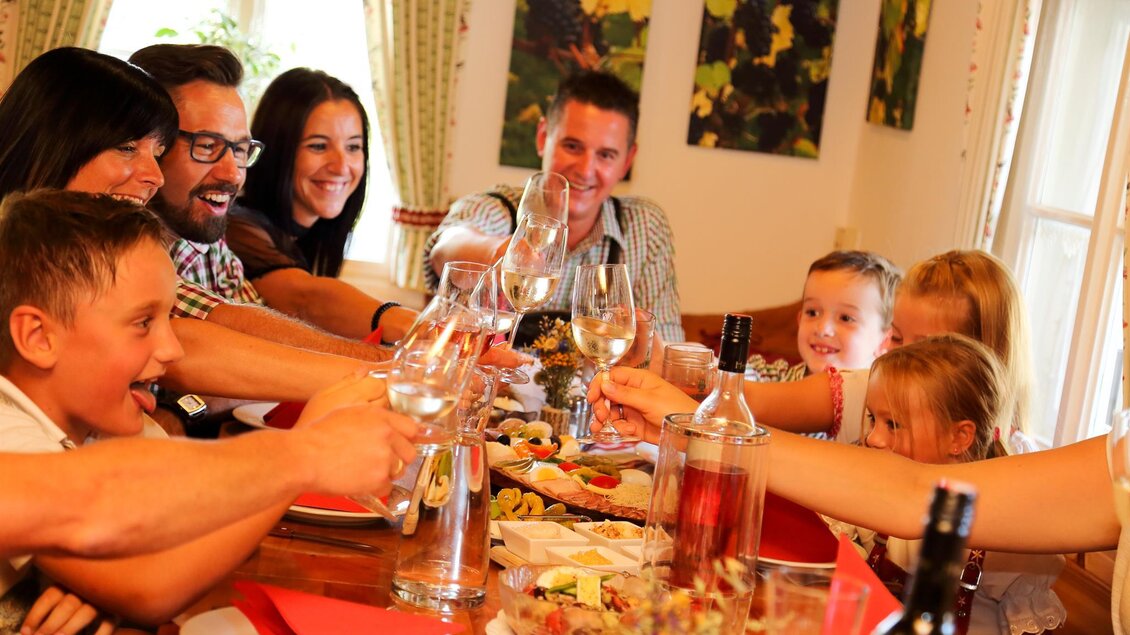 Eine fröhliche Zusammenkunft von Familienmitgliedern am Tisch. Sie stoßen mit Gläsern auf ein leckeres Essen an. | © Buschenschank Haubensima