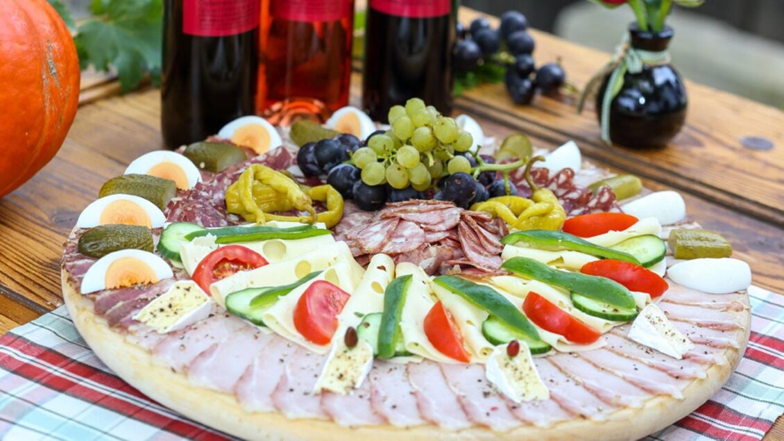 Eine üppige Wurst- und Käseplatte mit frischem Gemüse, Trauben und eingelegten Gurken. Im Hintergrund sind zwei Flaschen Wein zu sehen. | © Buschenschank Haubensima