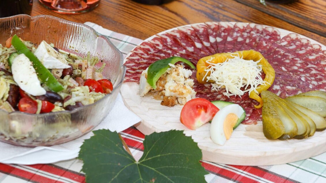 Ein Teller mit Wurst und Käse, garniert mit Paprika und Gurken, steht neben einem frischen Salat. Beide Gerichte liegen auf einem Holzbrett und sind an einem gedeckten Tisch angerichtet. | © Buschenschank Haubensima