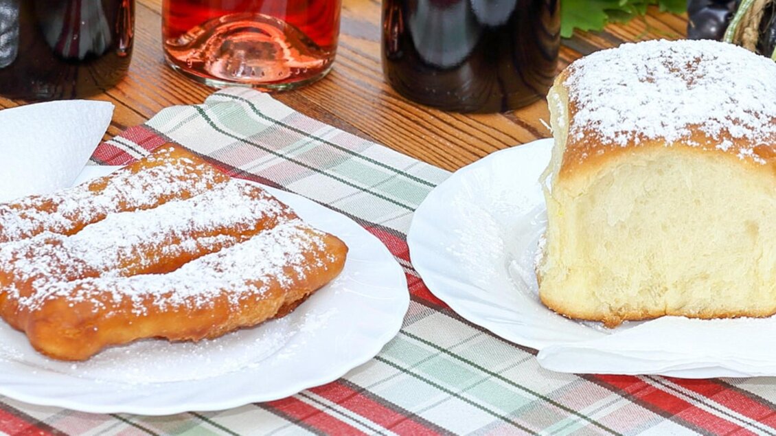 Ein Stück frittierter Teig mit Puderzucker und ein frisches, süßes Brot auf Tellern. Im Hintergrund sind einige Flaschen und eine kariert bedeckte Fläche zu sehen. | © Buschenschank Haubensima