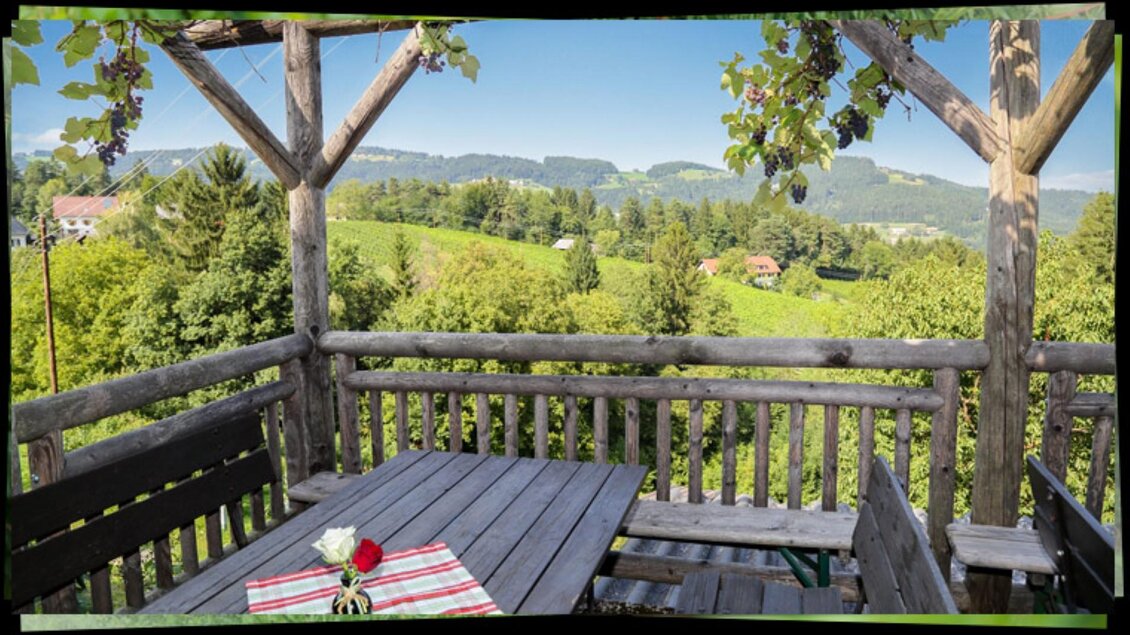 Ein schöner Balkon mit einem Holzgeländer und einem Tisch. Im Hintergrund erstrecken sich grüne Hügel und ein klarer Himmel. | © Buschenschank Haubensima