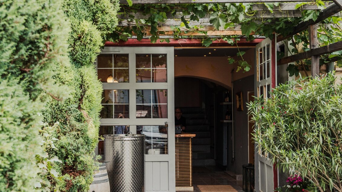 Ein einladender Eingang zu einem Restaurant, umgeben von grünen Pflanzen. Die Türen aus Holz sind geöffnet und vermitteln eine freundliche Atmosphäre. | © Buschenschank Hildebrand