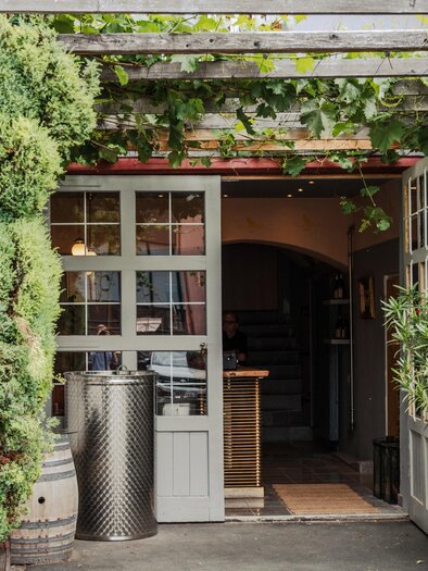 An inviting entrance to a restaurant, surrounded by green plants. The wooden doors are open, conveying a friendly atmosphere. | © Buschenschank Hildebrand