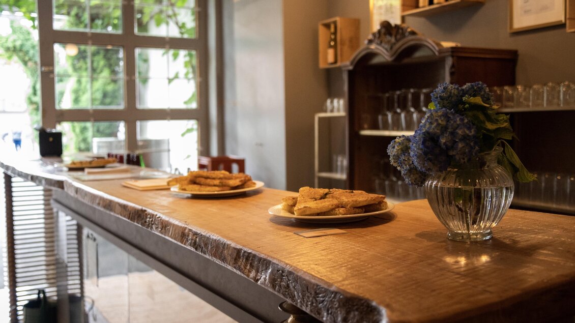 Eine einladende Café-Umgebung mit einem Holztheke. Auf dem Tisch stehen Gebäck und eine Vase mit blauen Blumen. | © Buschenschank Hildebrand